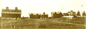 Black and white view across bare fields towards five buildings including an Italianate house and several barns.