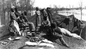 a ragtag looking group of black men, women and children with a few belongings standing on a levy by the water.