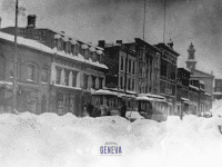 A commercial street of buildings with a trolley running down the middle and a man with a shovel and two horses behind large snowbanks.