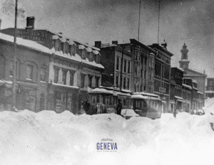 A commercial street of buildings with a trolley running down the middle and a man with a shovel and two horses behind large snowbanks.