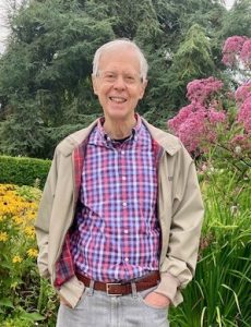 An older white-haired man in jeans, a plaid shirt, and beige jacket standing in a garden.