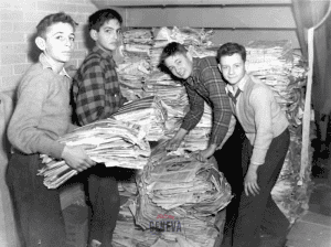 Four boys stacking piles of newspaper under a staircase.