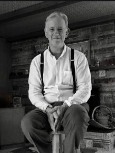 An older man in pants, white shirt and suspenders sitting on a stool in front of a stone fireplace.