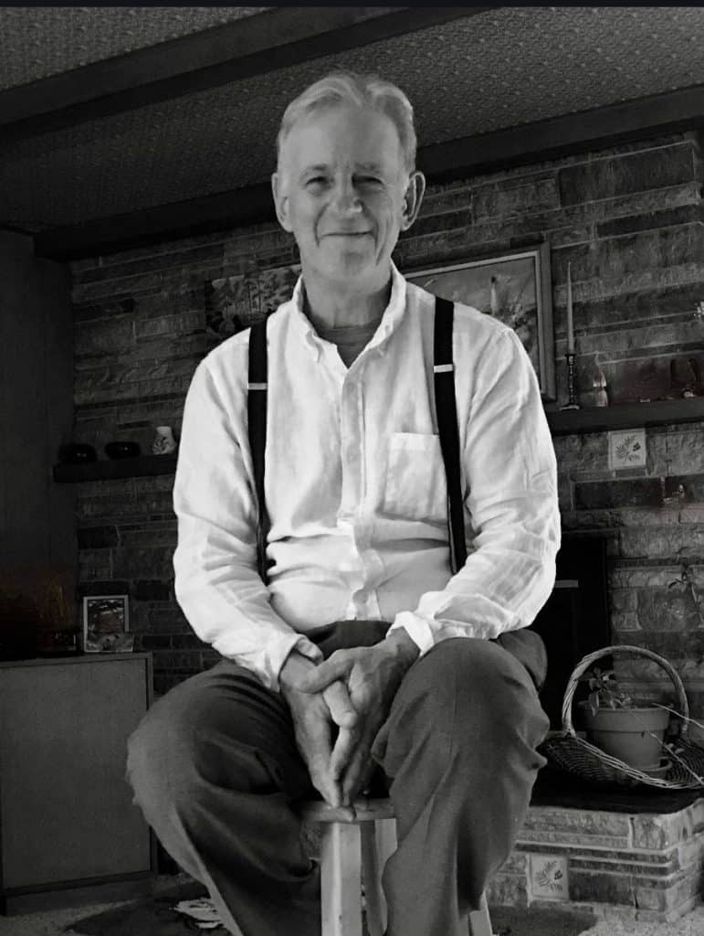 An older man in pants, white shirt and suspenders sitting on a stool in front of a stone fireplace.