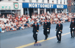 Uniformed firemen walk down a street lined with people sitting on the sidewalk in front of a Montgomery Ward store.