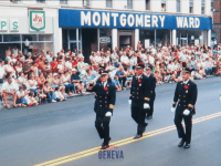 Uniformed firemen walk down a street lined with people sitting on the sidewalk in front of a Montgomery Ward store.