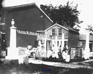 The yard outside Geneva Marble Works where a group of men in aprons and work clothes stand among the monuments and gravestones there.