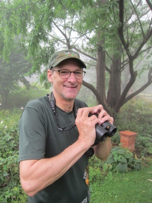 A man outside in a green bill cap and t-shirt with a pair of binoculars.