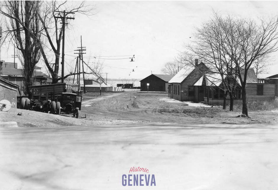 View down a dirt road with low buildings along it toward a lake.